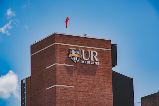 A tall brick building with the letters UR Medicine prominently displayed on it. A red flag is flying on top of the building under a bright blue sky with a few clouds.