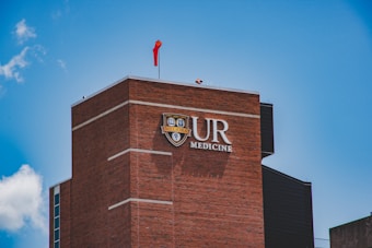 A tall brick building with the letters UR Medicine prominently displayed on it. A red flag is flying on top of the building under a bright blue sky with a few clouds.
