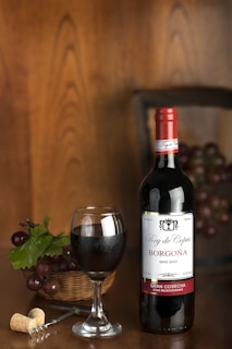 A bottle of red wine labeled 'Rey de Copas Borgoña' stands upright next to a filled wine glass. A basket of grapes with green leaves is placed nearby, along with a corkscrew and a cork. The background features a wooden texture, creating a warm ambiance.