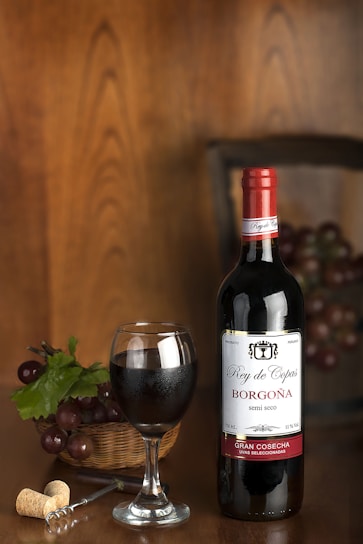 A bottle of red wine labeled 'Rey de Copas Borgoña' stands upright next to a filled wine glass. A basket of grapes with green leaves is placed nearby, along with a corkscrew and a cork. The background features a wooden texture, creating a warm ambiance.