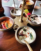 A wooden table with a brass stand holding multiple metal bowls containing a variety of colorful Indian dishes, including a white coconut-based chutney garnished with spices, red chili peppers, and curry leaves. A bright red flower is placed near the setup, adding a decorative touch.