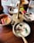 A wooden table with a brass stand holding multiple metal bowls containing a variety of colorful Indian dishes, including a white coconut-based chutney garnished with spices, red chili peppers, and curry leaves. A bright red flower is placed near the setup, adding a decorative touch.