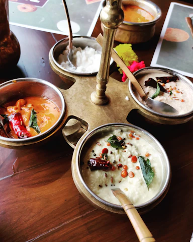 A wooden table with a brass stand holding multiple metal bowls containing a variety of colorful Indian dishes, including a white coconut-based chutney garnished with spices, red chili peppers, and curry leaves. A bright red flower is placed near the setup, adding a decorative touch.