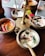 A wooden table with a brass stand holding multiple metal bowls containing a variety of colorful Indian dishes, including a white coconut-based chutney garnished with spices, red chili peppers, and curry leaves. A bright red flower is placed near the setup, adding a decorative touch.