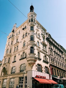 A tall, ornate building with an intricate architectural design stands against a clear blue sky. The building features numerous windows, balconies with decorative railings, and a turret at the top. A red awning with 'Monika' written on it in white letters is visible at street level, beside a few shop entrances. Overhead wires stretch across the sky.