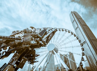 A futuristic sculpture resembling a mechanical dragon dominates the foreground. Behind it, a large Ferris wheel stands amidst a cluster of skyscrapers. The sky is overcast with scattered clouds, adding an industrial and urban feel to the scene.