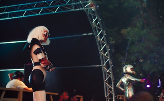 A person with a futuristic look stands in profile, wearing a black and white space-themed costume with a white wig and large sunglasses. Nearby, another individual in a metallic bodysuit and helmet appears to be pointing or gesturing. The setting includes a metal truss structure, and it's likely part of a performance or event.