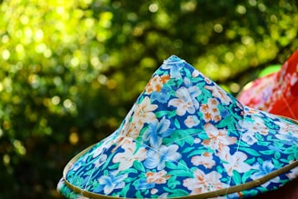 A colorful, floral-patterned conical hat set against a blurred green background. The pattern consists of blues, oranges, and whites with leaf and flower motifs. The background suggests a natural, outdoor setting with a bokeh effect.