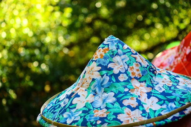 A colorful, floral-patterned conical hat set against a blurred green background. The pattern consists of blues, oranges, and whites with leaf and flower motifs. The background suggests a natural, outdoor setting with a bokeh effect.