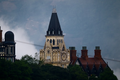 A gothic-style clock tower with intricate architectural details stands prominently. The building features a steep, dark slate roof and ornate stonework. Surrounding it are other buildings with similar architectural designs set against a foggy or misty backdrop, enhancing the mysterious ambiance.
