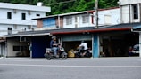 A man is riding a scooter along an urban street in an area with small shops or garages. Another person is visible in the background also riding a motorcycle. The buildings are simple, featuring different colored shutters and a variety of materials, such as brick and metal. Power lines crisscross above, indicating a typical street scene.