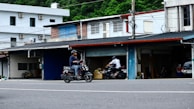 A man is riding a scooter along an urban street in an area with small shops or garages. Another person is visible in the background also riding a motorcycle. The buildings are simple, featuring different colored shutters and a variety of materials, such as brick and metal. Power lines crisscross above, indicating a typical street scene.