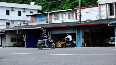 A man is riding a scooter along an urban street in an area with small shops or garages. Another person is visible in the background also riding a motorcycle. The buildings are simple, featuring different colored shutters and a variety of materials, such as brick and metal. Power lines crisscross above, indicating a typical street scene.