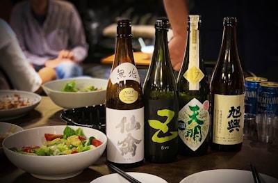 Several bottles of sake are lined up on a table next to dishes of food, including a bowl of salad. There are also some empty plastic cups and beer cans in the background. People are seated around the table engaging in a social gathering.