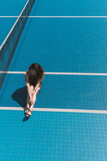 A woman is sitting on a blue tennis court near the net. She appears relaxed and contemplative with her hands wrapped around her knees. The court is brightly lit by the sun, creating a sharp shadow next to her.