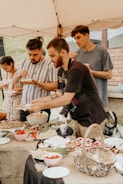 A group of people enjoying a meal at an outdoor gathering under a tent. Various dishes, including vegetables and bread, are laid out on a table covered with a burlap cloth. The individuals are casually dressed, with one person holding a camera.