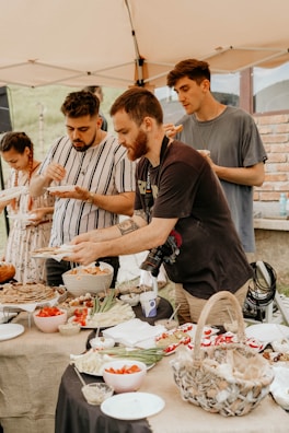 A group of people enjoying a meal at an outdoor gathering under a tent. Various dishes, including vegetables and bread, are laid out on a table covered with a burlap cloth. The individuals are casually dressed, with one person holding a camera.