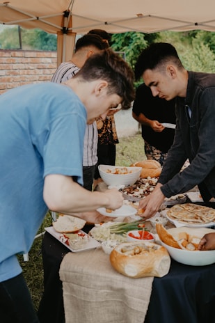A group of people are gathered around a table set with various dishes, including bread, vegetables, dips, and grilled items. The scene takes place under a canopy outdoors, with greenery and a brick wall in the background.