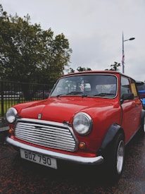 A classic red Mini Cooper car is parked outdoors on a wet pavement. The vintage vehicle features a chrome grille and headlights, with a license plate displaying 'BDZ 790'. Trees and a street lamp are visible in the background, along with a Union Jack flag on a pole.