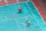 A person in red shorts is cleaning or working on a weathered, turquoise sports court. The court has visible wear and patches of different shades. There is a chair with objects including bottles placed on the ground. A broom is also visible.