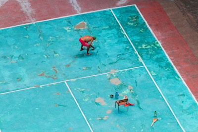 A person in red shorts is cleaning or working on a weathered, turquoise sports court. The court has visible wear and patches of different shades. There is a chair with objects including bottles placed on the ground. A broom is also visible.