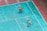 A person in red shorts is cleaning or working on a weathered, turquoise sports court. The court has visible wear and patches of different shades. There is a chair with objects including bottles placed on the ground. A broom is also visible.