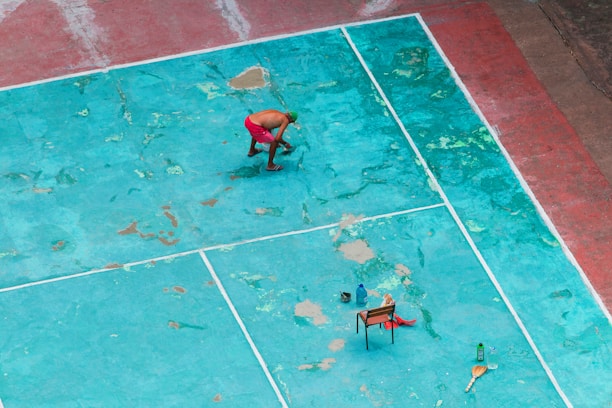 A person in red shorts is cleaning or working on a weathered, turquoise sports court. The court has visible wear and patches of different shades. There is a chair with objects including bottles placed on the ground. A broom is also visible.