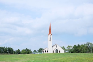 A white church with a tall red steeple is situated in a lush green field. Surrounding the church are several trees, and above, a blue sky with scattered white clouds creates a serene atmosphere.