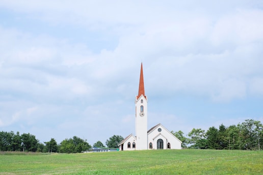 A white church with a tall red steeple is situated in a lush green field. Surrounding the church are several trees, and above, a blue sky with scattered white clouds creates a serene atmosphere.