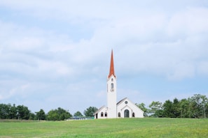A white church with a tall red steeple is situated in a lush green field. Surrounding the church are several trees, and above, a blue sky with scattered white clouds creates a serene atmosphere.