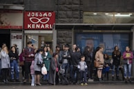 A group of people stand and wait at a bus stop in an urban environment. The backdrop includes storefronts with signs in a language using a distinct script, possibly Georgian. People are dressed in various styles, suggesting a cool or mild climate.