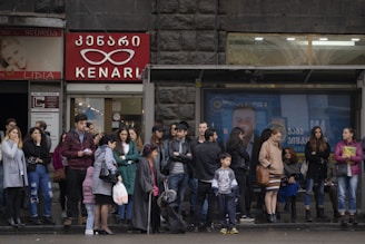 A group of people stand and wait at a bus stop in an urban environment. The backdrop includes storefronts with signs in a language using a distinct script, possibly Georgian. People are dressed in various styles, suggesting a cool or mild climate.