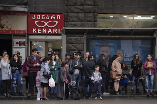 A group of people stand and wait at a bus stop in an urban environment. The backdrop includes storefronts with signs in a language using a distinct script, possibly Georgian. People are dressed in various styles, suggesting a cool or mild climate.