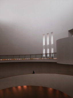 The image features a minimalist architectural interior with curved walls, large open spaces, and a few vertical slits for natural light. A solitary figure is seated on one of the curved ledges within the space, contributing to the serene and contemplative atmosphere.