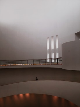 The image features a minimalist architectural interior with curved walls, large open spaces, and a few vertical slits for natural light. A solitary figure is seated on one of the curved ledges within the space, contributing to the serene and contemplative atmosphere.