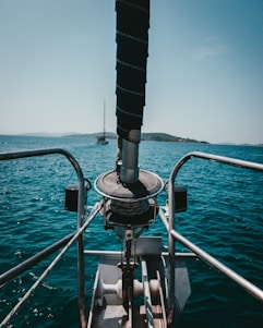 A boat bow with metallic railings and a winch system is visible against the backdrop of a calm blue sea and distant islands. A sailboat can be seen in the distance under a clear sky.