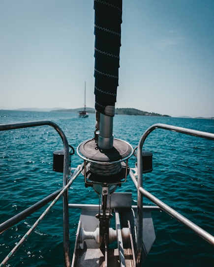 A boat bow with metallic railings and a winch system is visible against the backdrop of a calm blue sea and distant islands. A sailboat can be seen in the distance under a clear sky.