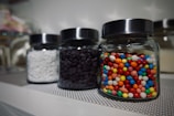 Three glass jars with dark lids are positioned on a table. The jar in the foreground contains a variety of colorful candies, including red, blue, yellow, and green. The second jar holds small dark chocolate chips, and the third jar contains small white candies or confections.