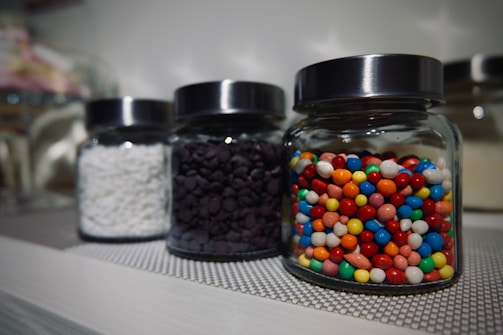 Three glass jars with dark lids are positioned on a table. The jar in the foreground contains a variety of colorful candies, including red, blue, yellow, and green. The second jar holds small dark chocolate chips, and the third jar contains small white candies or confections.