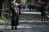 An elderly person wearing a colorful scarf and dark clothing is walking with a cane on a sunlit path lined with trees. Other people are visible in the distance, along with a red cafe umbrella and a child crouched down to the right.