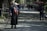 An elderly person wearing a colorful scarf and dark clothing is walking with a cane on a sunlit path lined with trees. Other people are visible in the distance, along with a red cafe umbrella and a child crouched down to the right.