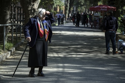 An elderly person wearing a colorful scarf and dark clothing is walking with a cane on a sunlit path lined with trees. Other people are visible in the distance, along with a red cafe umbrella and a child crouched down to the right.