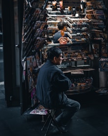 A person is sitting on a stool in front of a busy newsstand filled with magazines, newspapers, and snacks. The scene is dimly lit, creating a moody atmosphere, and the person appears to be absorbed in their surroundings.