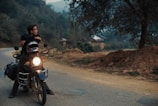 A person is sitting on a motorcycle parked on a rural road, gazing into the distance. The setting is serene, surrounded by trees and hills, with a soft, overcast sky. The motorcycle is equipped with luggage on the back, suggesting a journey or adventure. Dirt and foliage line the road, hinting at a remote or countryside location.