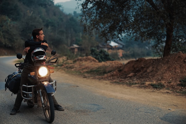 A person is sitting on a motorcycle parked on a rural road, gazing into the distance. The setting is serene, surrounded by trees and hills, with a soft, overcast sky. The motorcycle is equipped with luggage on the back, suggesting a journey or adventure. Dirt and foliage line the road, hinting at a remote or countryside location.