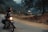 A person is sitting on a motorcycle parked on a rural road, gazing into the distance. The setting is serene, surrounded by trees and hills, with a soft, overcast sky. The motorcycle is equipped with luggage on the back, suggesting a journey or adventure. Dirt and foliage line the road, hinting at a remote or countryside location.