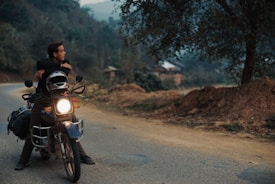 A person is sitting on a motorcycle parked on a rural road, gazing into the distance. The setting is serene, surrounded by trees and hills, with a soft, overcast sky. The motorcycle is equipped with luggage on the back, suggesting a journey or adventure. Dirt and foliage line the road, hinting at a remote or countryside location.