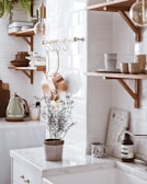 A cozy kitchen scene features wooden shelves with white and brown dishes, copper pots hanging from a rod, and a rustic kettle on a counter. A plant in a pot sits on a white marble countertop, accompanied by a patterned mug and a bottle. The background showcases subway tile walls, natural wood accents, and a soft, warm lighting.