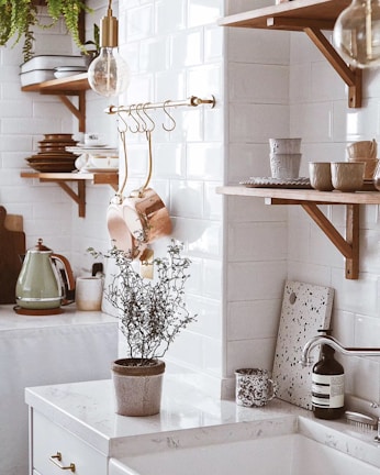 A cozy kitchen scene features wooden shelves with white and brown dishes, copper pots hanging from a rod, and a rustic kettle on a counter. A plant in a pot sits on a white marble countertop, accompanied by a patterned mug and a bottle. The background showcases subway tile walls, natural wood accents, and a soft, warm lighting.