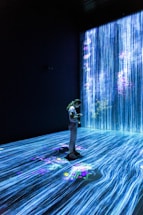 A person stands in a dark room surrounded by digital projections of cascading blue vertical lines resembling a waterfall, with colorful floral patterns scattered throughout. The room is illuminated by the vivid projections, creating an immersive and serene environment.